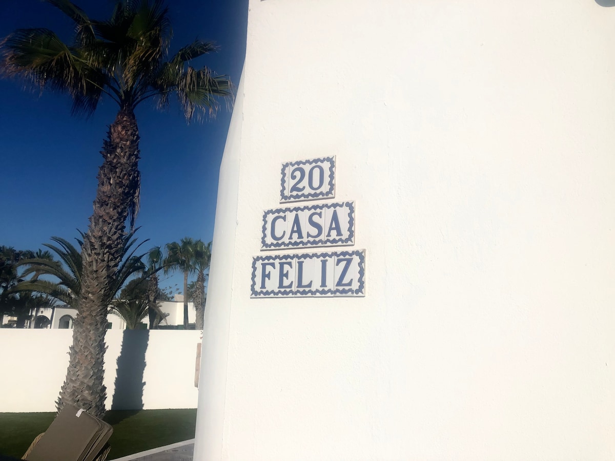 A white wall displays the villa's address, '20 CASA FELIZ,' highlighted with decorative tiles. Palm trees are visible in the background, contributing to the villa's tropical setting.