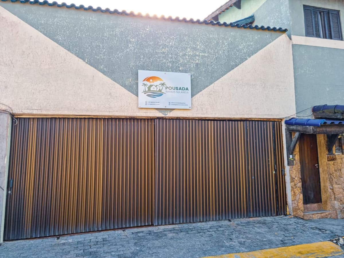 The exterior of the building is shown, featuring a large metal rolling gate that is closed. A sign identifying the property is visible above the gate. The structure displays a blend of textured walls and a tiled roof, framed by simple architectural lines.