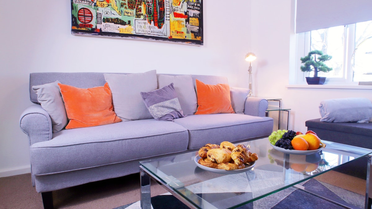 A modern living room is displayed featuring a comfortable gray sofa adorned with orange and purple cushions. A glass coffee table holds a plate of pastries, complemented by a bowl of fresh fruit. Colorful wall art adds a lively touch, and natural light streams through a nearby window.