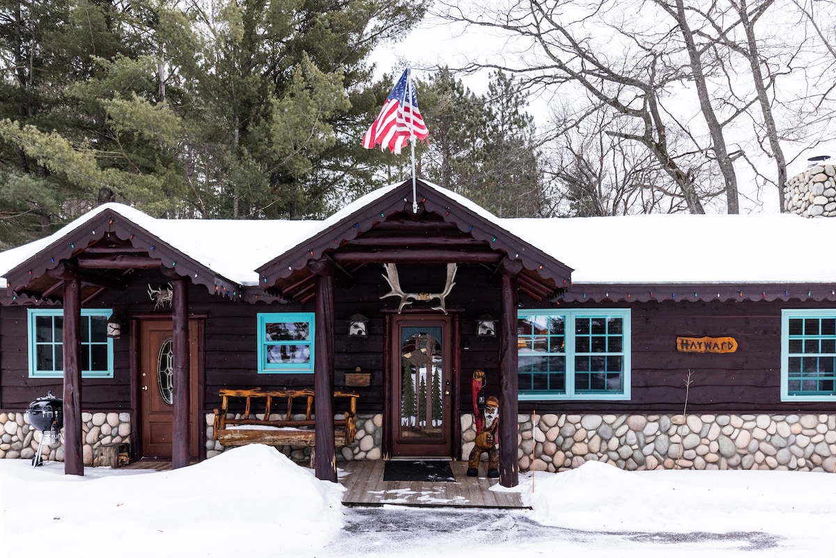The cabin's exterior features dark wooden siding, accented by bright blue windows. An American flag flies above the entrance, framed by snow-covered grounds. Unique decorations, including antlers and a stone facade, enhance the rustic charm of this cozy retreat.