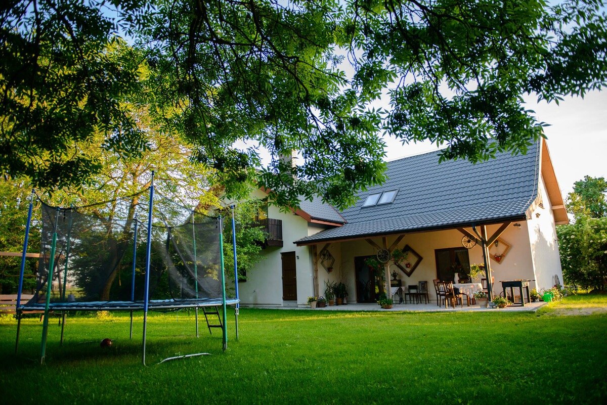 A spacious house is set in a serene garden, with a large trampoline situated on the green lawn. The exterior features a sloped roof and multiple windows, while a covered patio holds a table and chairs, surrounded by lush trees.