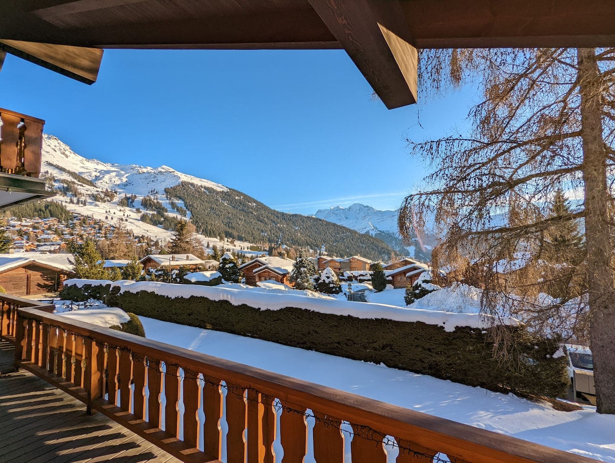 A clear view of snow-covered mountains is framed by wooden railings on the terrace. The landscape features a mix of chalets nestled among evergreen trees, with a bright blue sky visible above. Snow blankets the ground, creating a serene winter scene.