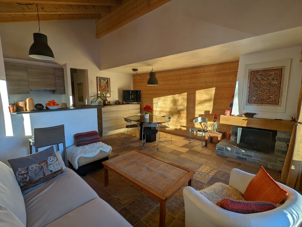A spacious living area is shown with a light-colored sofa and small armchairs. A wooden coffee table sits in the center. The kitchen is partially visible, equipped with modern cabinetry. Warm sunlight filters through, highlighting the textured stone flooring and wooden accent wall.