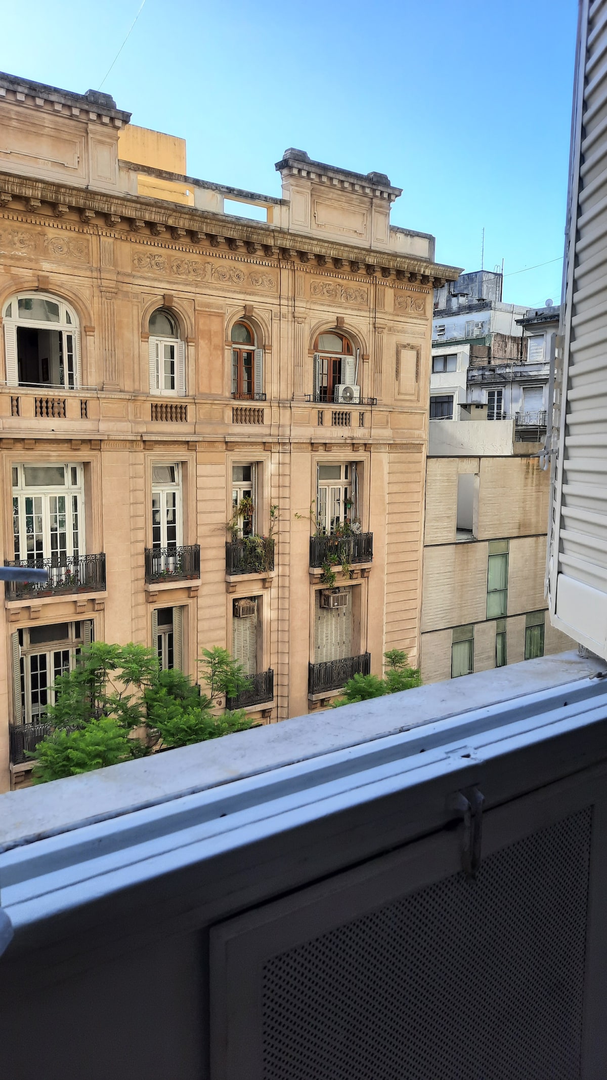 A view from the window captures a historic building with intricate architectural details and balconies adorned with greenery. The blue sky provides a bright backdrop, enhancing the visual appeal of the urban scenery.