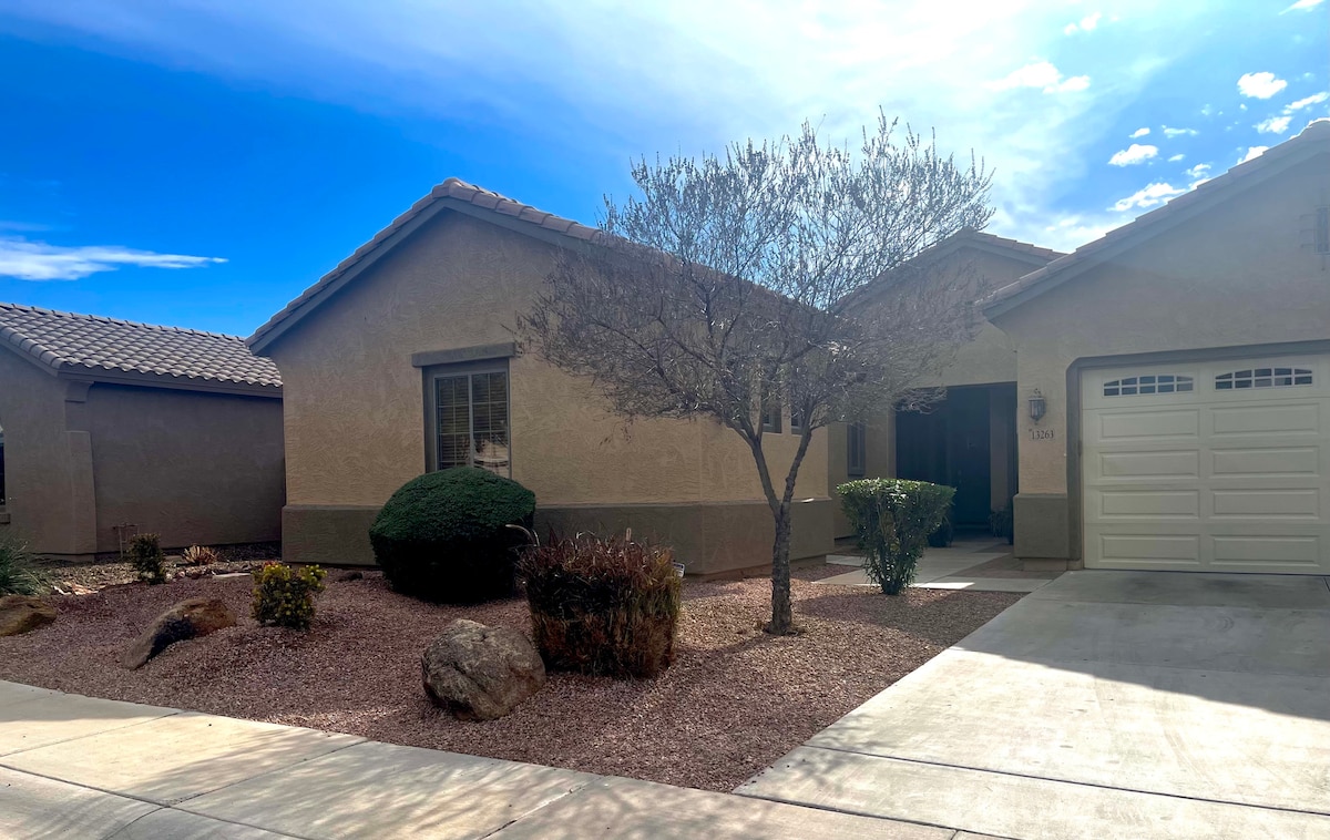 The exterior of the guest suite features a single-story structure with a beige stucco finish and a tiled roof. A manicured front yard includes a small tree and various plants, with gravel landscaping and a driveway leading to a garage.