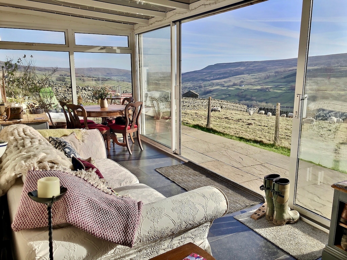 A bright conservatory features large glass doors offering expansive views of the surrounding valley. Comfortably furnished with a light-colored settee and a dining table, the space invites relaxation while enjoying the natural scenery. A textured throw and decorative cushions add comfort to the seating area.