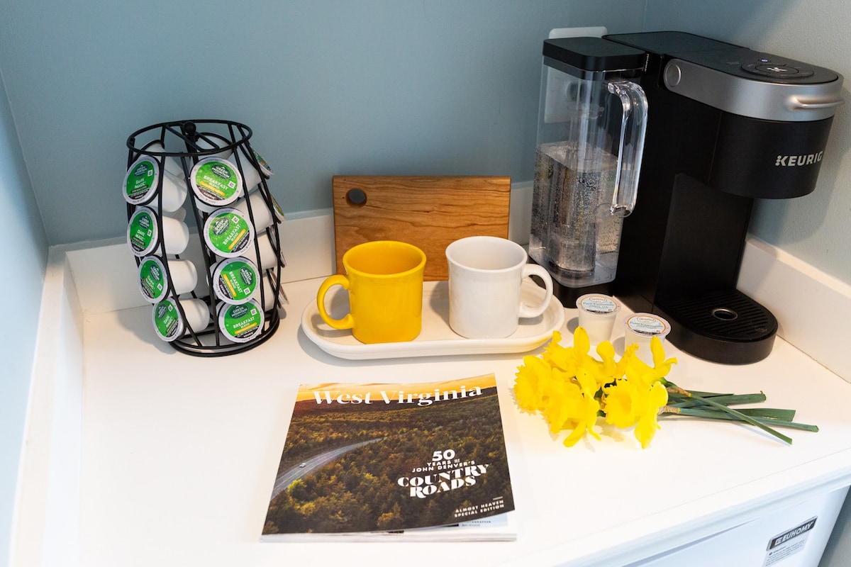 A kitchenette area is presented, featuring a Keurig coffee maker alongside a variety of coffee pods. Two cups are positioned on a tray, one yellow and one white. A wooden cutting board and a magazine titled 'West Virginia: 50 Must-See Scenic Country Roads' are also included, with a small vase of yellow flowers.
