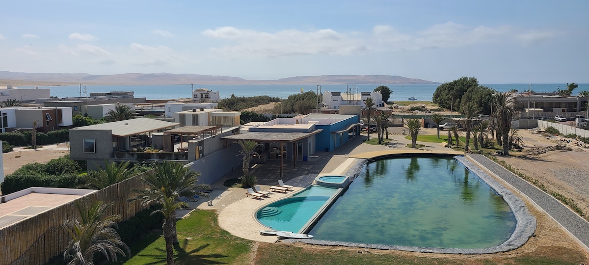 An aerial view captures the expansive grounds of the property, featuring a large, vibrant green saltwater lagoon and a separate pool. The landscape includes palm trees and modern structures, with Paracas Bay visible in the background under a clear sky.