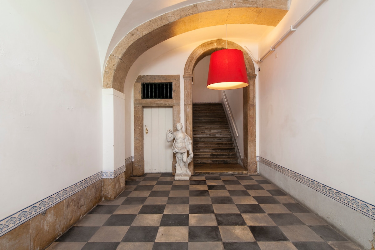 A spacious entryway is highlighted by a checkered tile floor and an arched ceiling. A white statue is positioned centrally, and a large red lampshade hangs from above, drawing attention. A staircase is visible, leading to the upper levels.