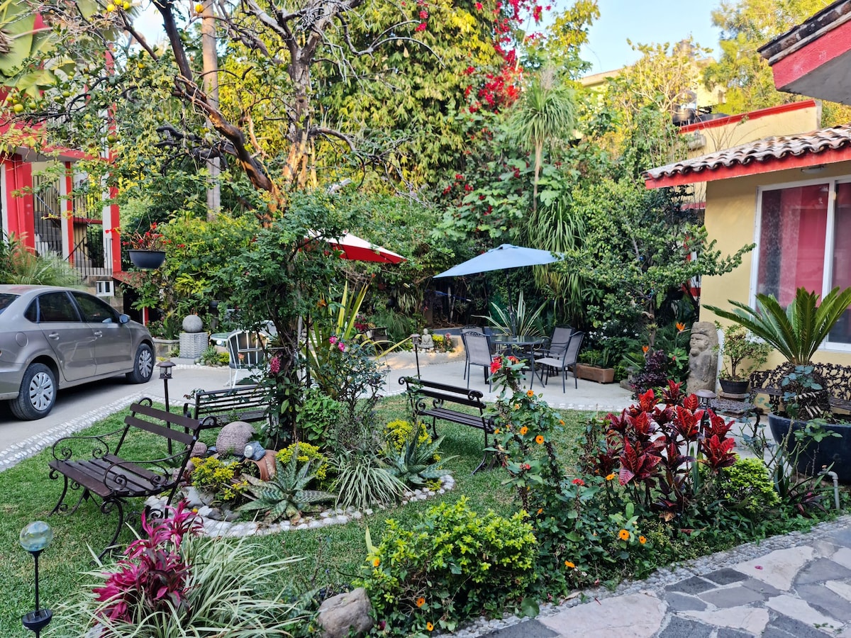 A lush garden area is presented, featuring a variety of plants, flowers, and decorative stones. Seating options include benches and umbrellas, providing shaded spots for relaxation. The vibrant greenery is complemented by a stone pathway and glimpses of nearby buildings, enhancing the tranquil environment.