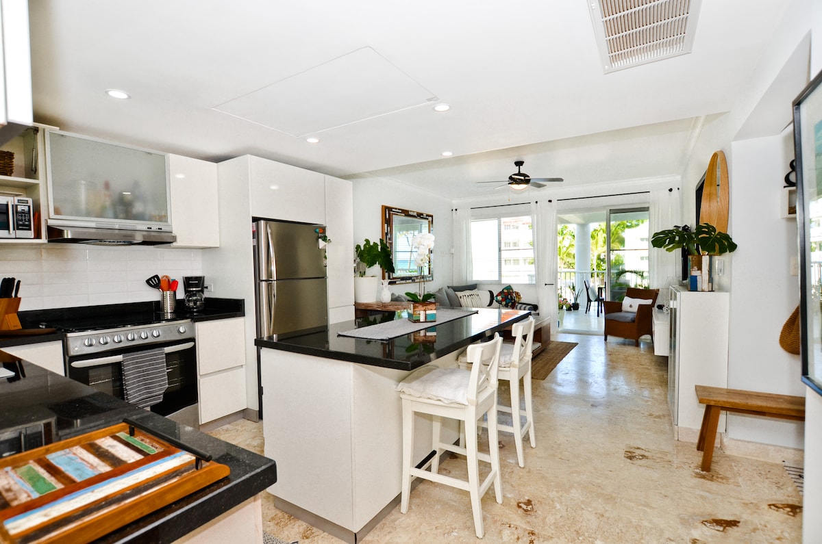 A modern kitchen is showcased, featuring sleek white cabinetry and a large central island with seating. Stainless steel appliances, including a refrigerator and oven, are visible. Natural light floods the space through nearby windows that lead to a living area with comfortable seating.