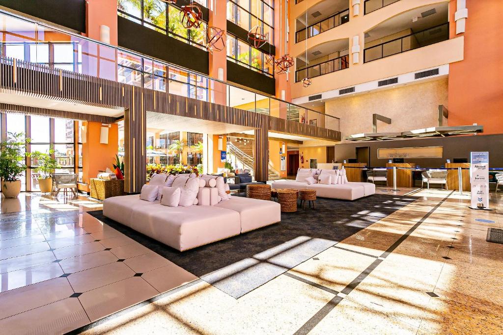 A spacious lobby area is highlighted by large windows allowing natural light to fill the space. Comfortable white seating areas are arranged around wooden tables, creating inviting spaces for relaxation. Green plants add a touch of nature to the contemporary design.