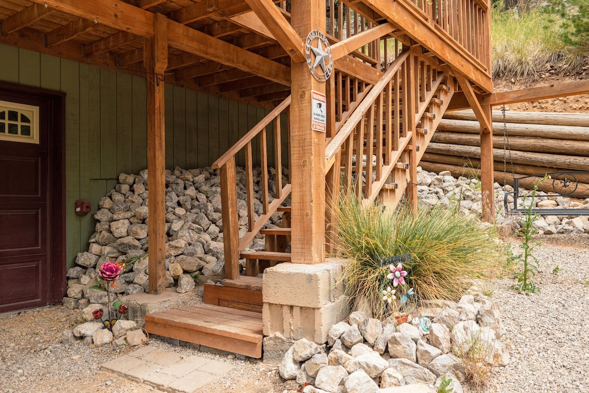 The entrance features a wooden staircase leading to the upper level. Natural stone landscaping surrounds the base, enhanced by decorative flowers. A single door is visible to the left, framed by wooden beams of the structure above.