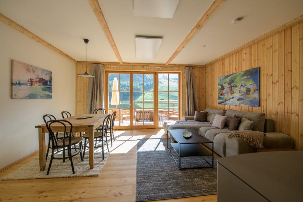 A spacious living area is showcased, featuring a comfortable gray sofa and a wooden dining table with black chairs. Large windows provide views of the surrounding nature, while natural light brightens the wooden walls and flooring. Art pieces adorn the walls, adding color to the setting.