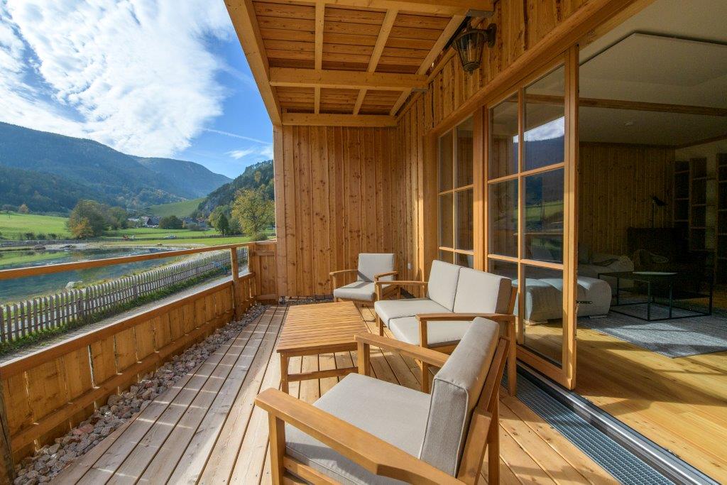 A wooden terrace features comfortable seating with light-colored cushions and a wooden table, overlooking lush green landscapes and hills. Large glass doors provide access to the interior space, while the natural wood paneling adds warmth to the setting.