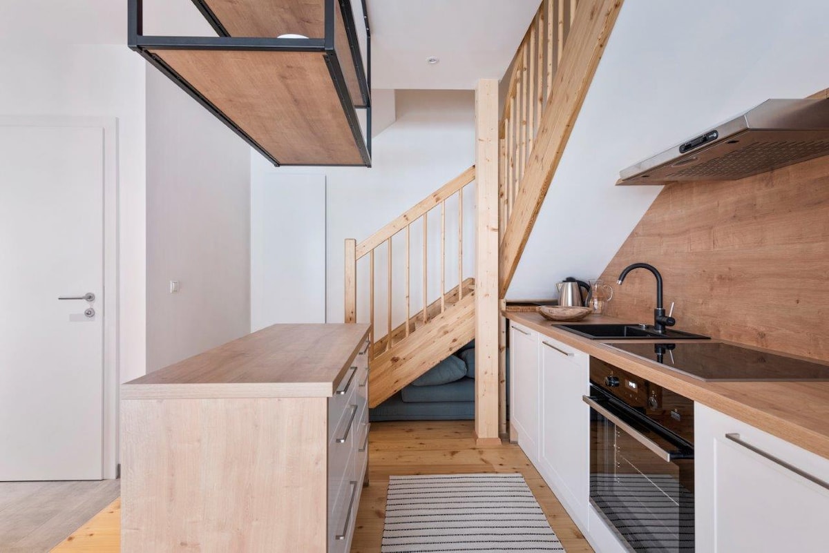 A modern kitchen features wooden cabinetry and a central island with a sleek countertop. A stairway with wooden steps is positioned in the background. Appliances include an oven and a kettle, with a rug laid on the wooden floor.