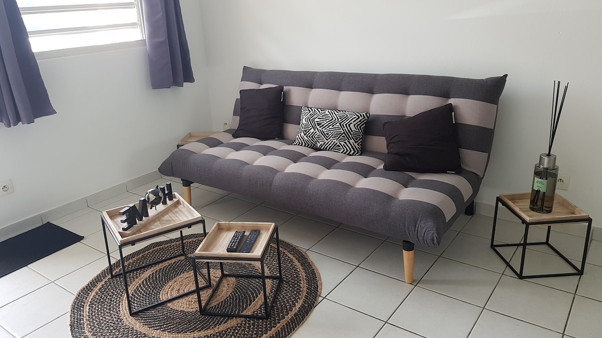 A modern seating area features a striped futon with dark throw pillows. Two small, rectangular tables are positioned beside the futon, one displaying decorative items. A round woven rug adds texture to the tiled floor, while light filters through window treatments in a neutral tone.