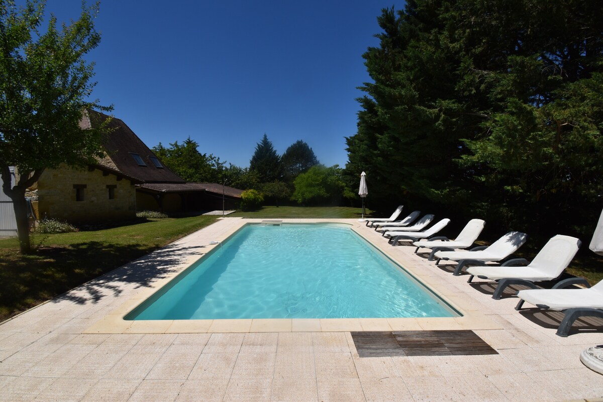 A heated swimming pool is surrounded by a sun-drenched terrace with several lounge chairs for relaxation. Lush greenery and trees frame the area, providing a serene outdoor setting. The well-maintained lawn is visible beyond the pool, enhancing the tranquil ambiance.