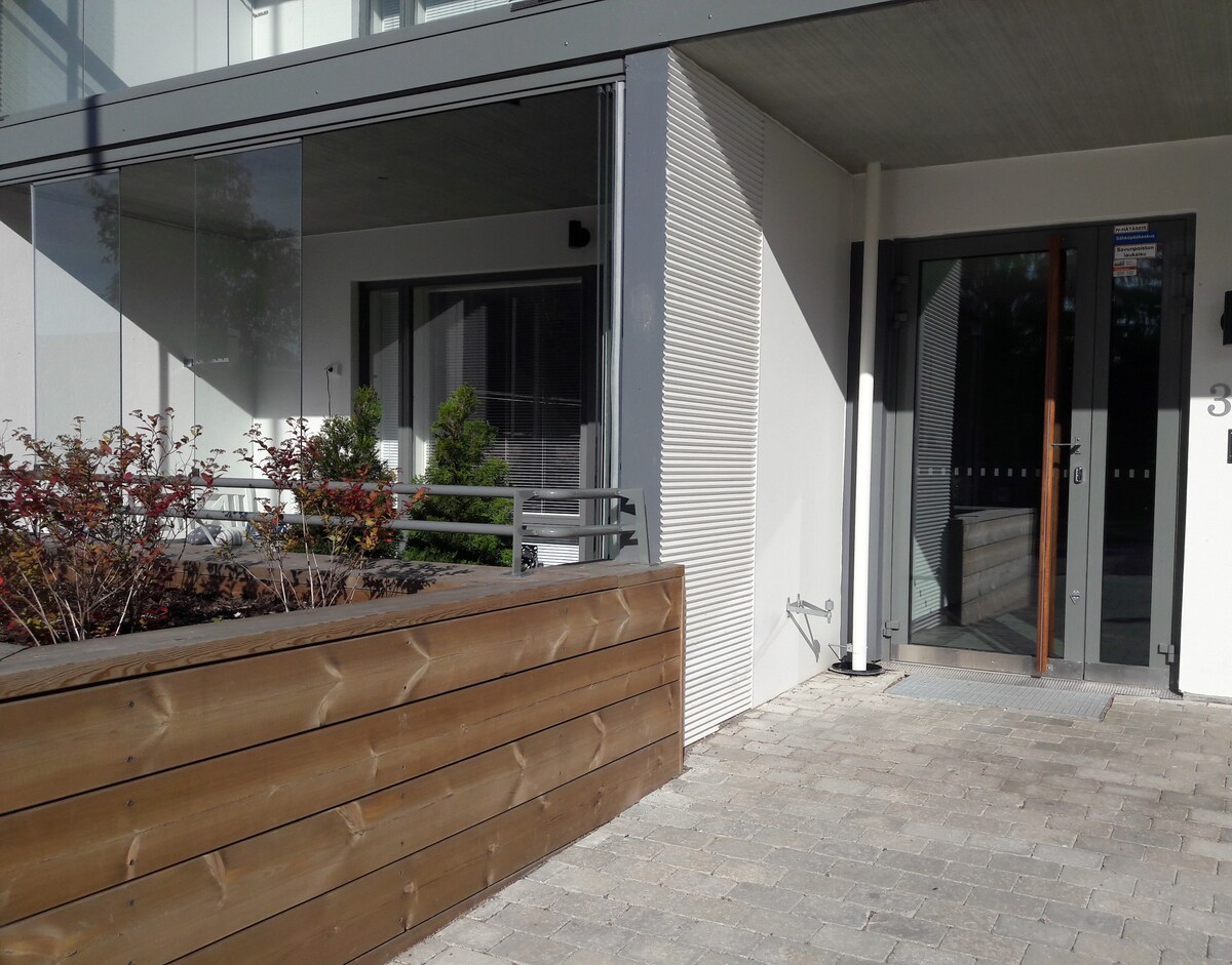 The entrance of the building is depicted, featuring a modern door with glass panels and a welcoming pathway. Surrounding greenery and a wooden planter add a touch of nature. Light reflects off the surfaces, suggesting a bright atmosphere.