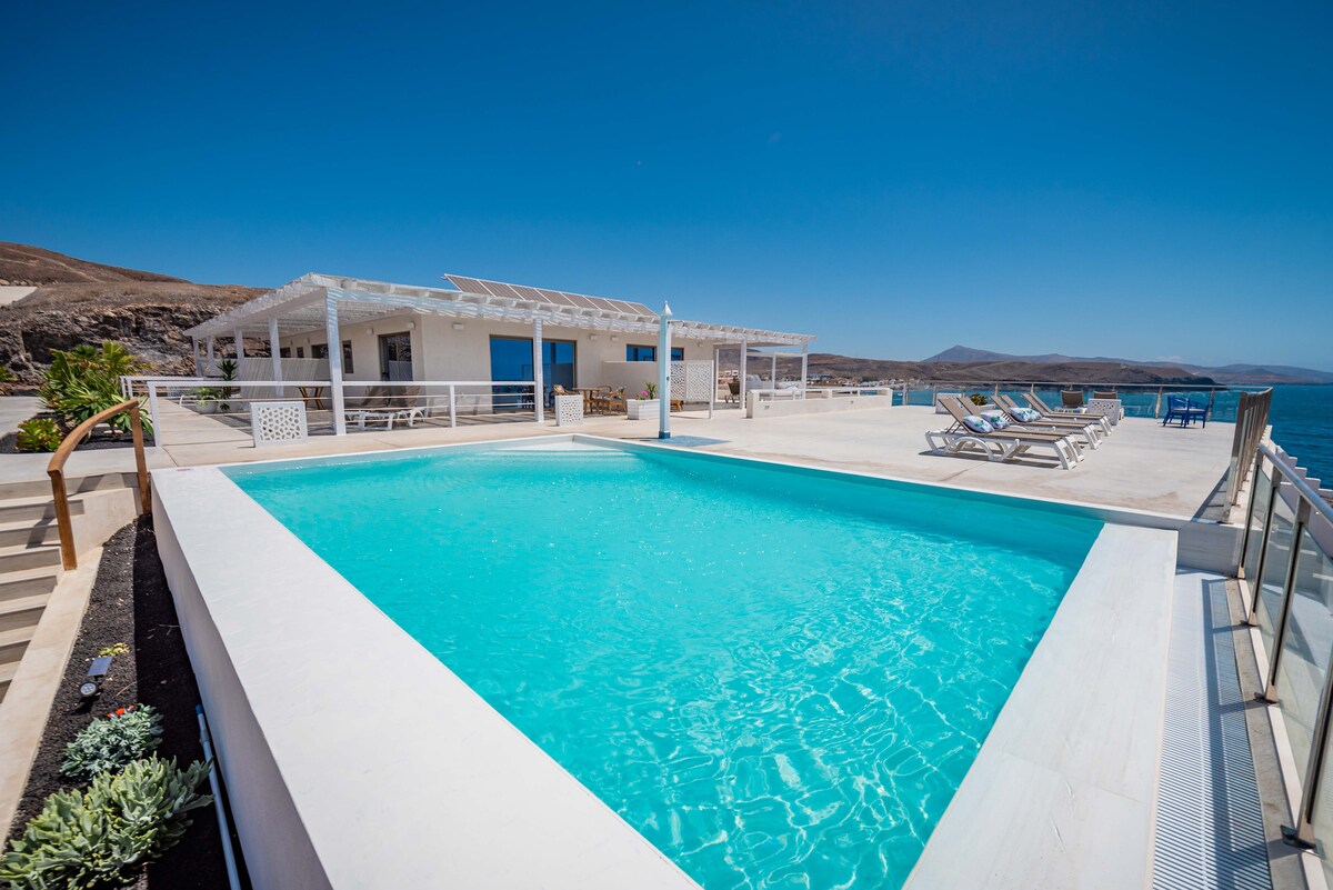 An inviting infinity pool with clear blue water is surrounded by a spacious terrace. Sun loungers are arranged along the edge, offering panoramic views of the ocean. A modern white building is visible in the background, complementing the serene coastal environment.