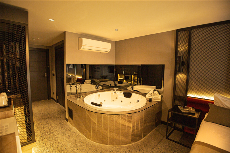 A modern bathroom is featured, showcasing a large, circular spa tub with jets. The space is highlighted by sleek, reflective surfaces and softly illuminated by ambient lighting. A vanity area with toiletries is situated nearby, complementing the contemporary design.