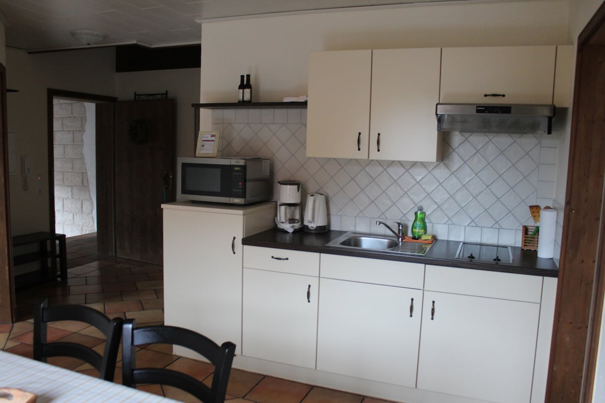 The kitchen features a compact layout with white cabinetry, a sink, and a small microwave. A coffee maker and kettle are visible on the counter. Tile flooring complements the space, with a dining table and chairs situated nearby.