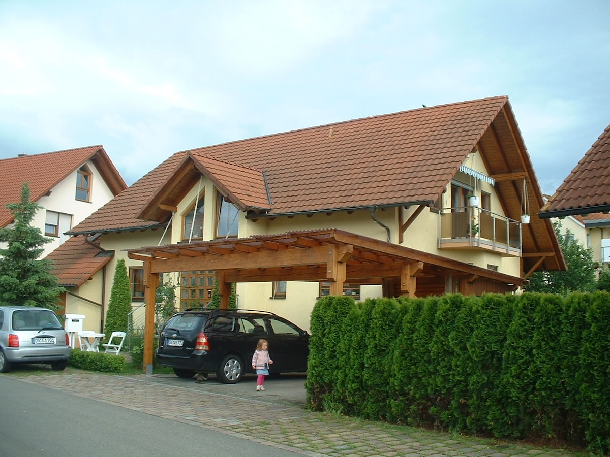 A two-story guest house with a steeply pitched roof and wooden accents is surrounded by neatly trimmed greenery. A car is parked under a wooden carport, and a child stands on the pathway, highlighting the welcoming outdoor space.