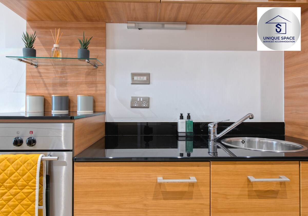 A modern kitchen space is featured with sleek wooden cabinetry and a black countertop. Stainless steel appliances, including an oven and a sink, are present. Small potted plants and neatly arranged toiletries add touches of greenery and organization to the countertop.