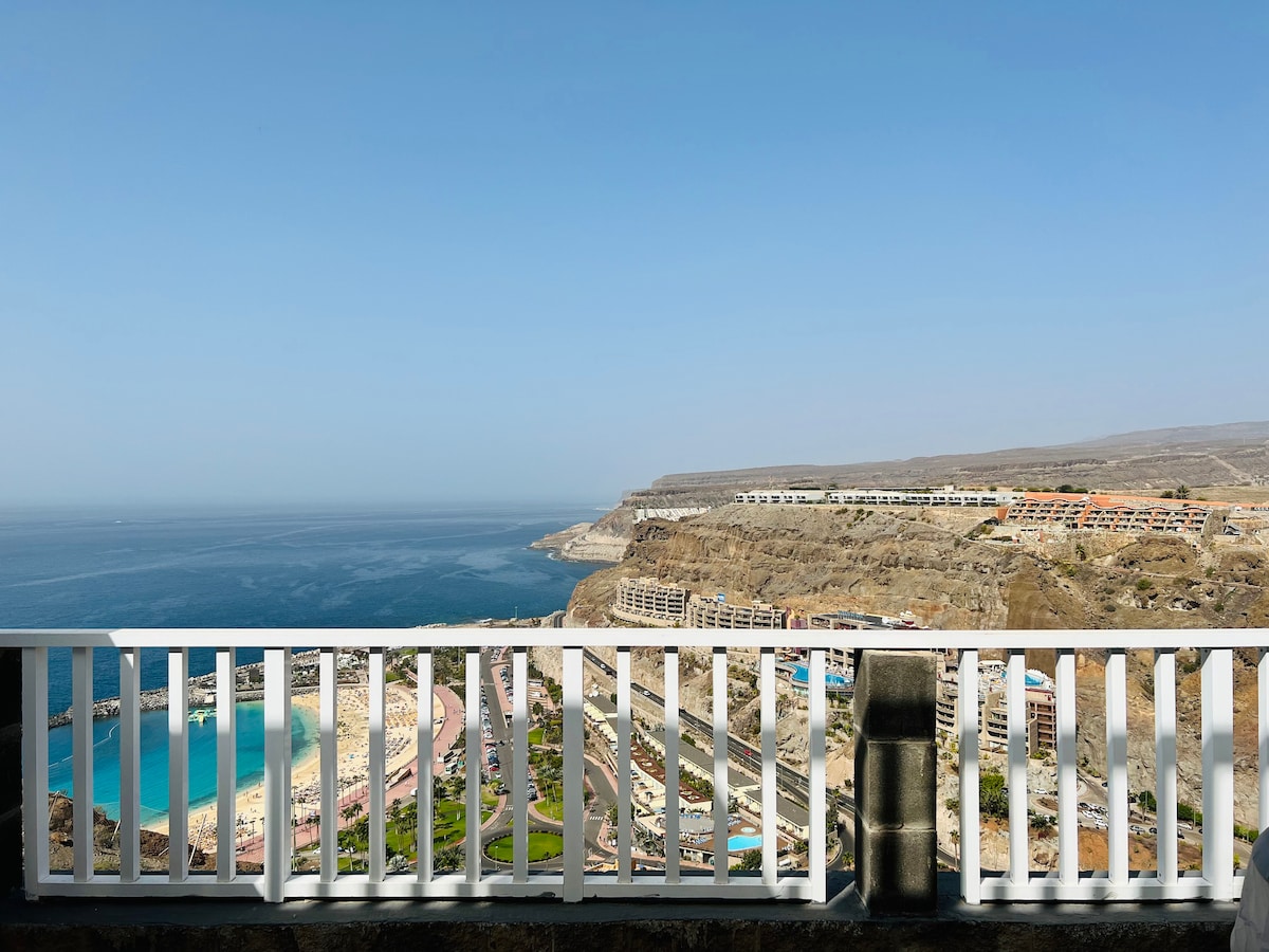 A panoramic view from a private terrace showcases the coastline and the ocean. The horizon features distant cliffs and buildings along the shoreline. A clear blue sky complements the vibrant turquoise waters, providing a serene atmosphere that invites relaxation.