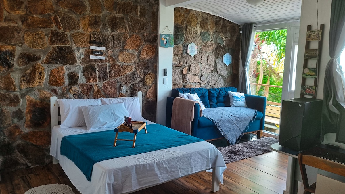 A cozy bedroom features a spacious bed with a blue cover and decorative pillows, complemented by a stone wall. A comfortable sofa in soft blue is positioned beside large windows that invite natural light and offer views of the outdoor greenery.