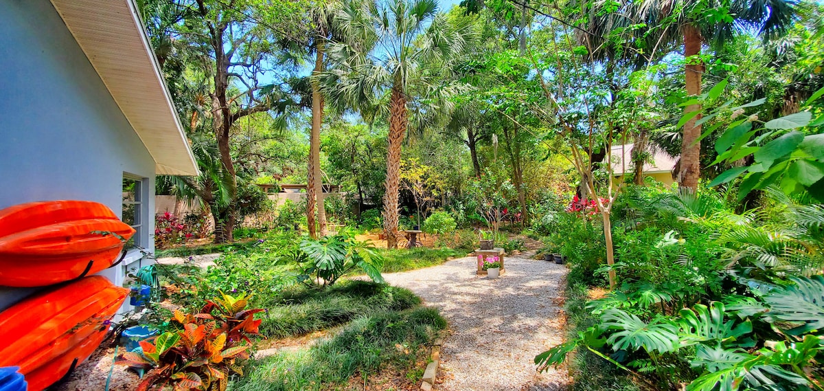 A lush garden is surrounded by towering palm trees and various native plants and flowers. A winding path of gravel leads through the greenery, with vibrant foliage and blooming flowers providing a natural habitat for local wildlife. Nearby, kayaks are stored against a wall.
