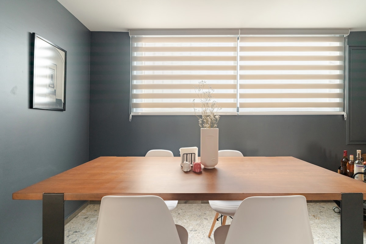 A modern dining area features a wooden table accompanied by white chairs. Natural light filters through two layered window treatments, creating a soft atmosphere. On the table, a vase holds dry foliage, while various bottles are arranged in the corner, adding subtle warmth to the space.