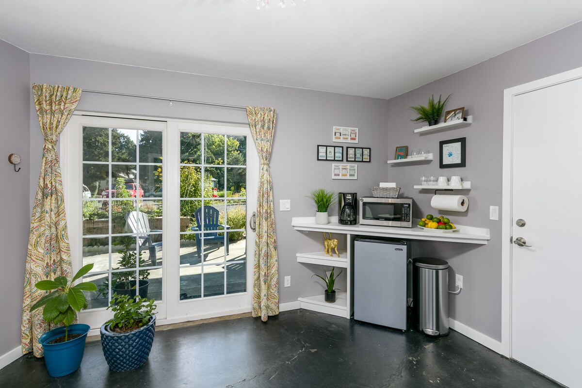 A well-appointed kitchenette area features a refrigerator, microwave, and coffee maker, set against a light grey wall. Decorative plants and dishes are arranged on open shelves. Sliding glass doors allow access to the outside garden, with vibrant colors highlighted by natural light.