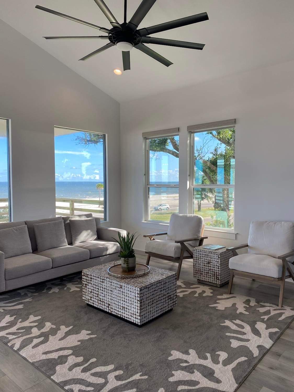 A bright living area features a gray sofa and two light-colored armchairs, arranged around a textured coffee table. Large windows frame views of the surroundings, allowing natural light to fill the space. A decorative rug with coral patterns adds a subtle touch.