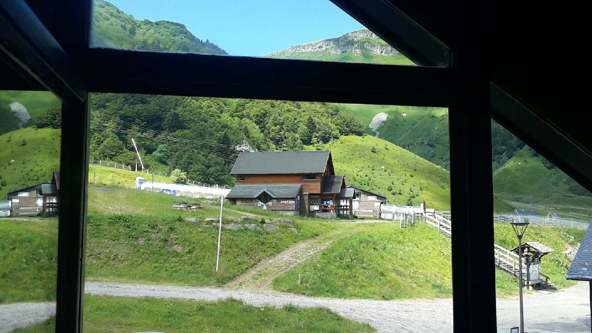 A view from the window showcases rolling green hills under a clear sky. A rustic building is visible in the distance, surrounded by vibrant grass. A gravel path leads towards the structure, enhancing the natural landscape.
