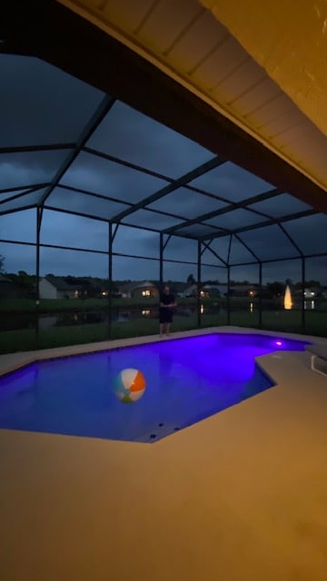 A private pool area is enclosed by a screen, featuring a colorful beach ball floating on the water's surface. Dim lighting casts a soft purple hue in the pool, set against a twilight sky and surrounding homes illuminated by distant lights.