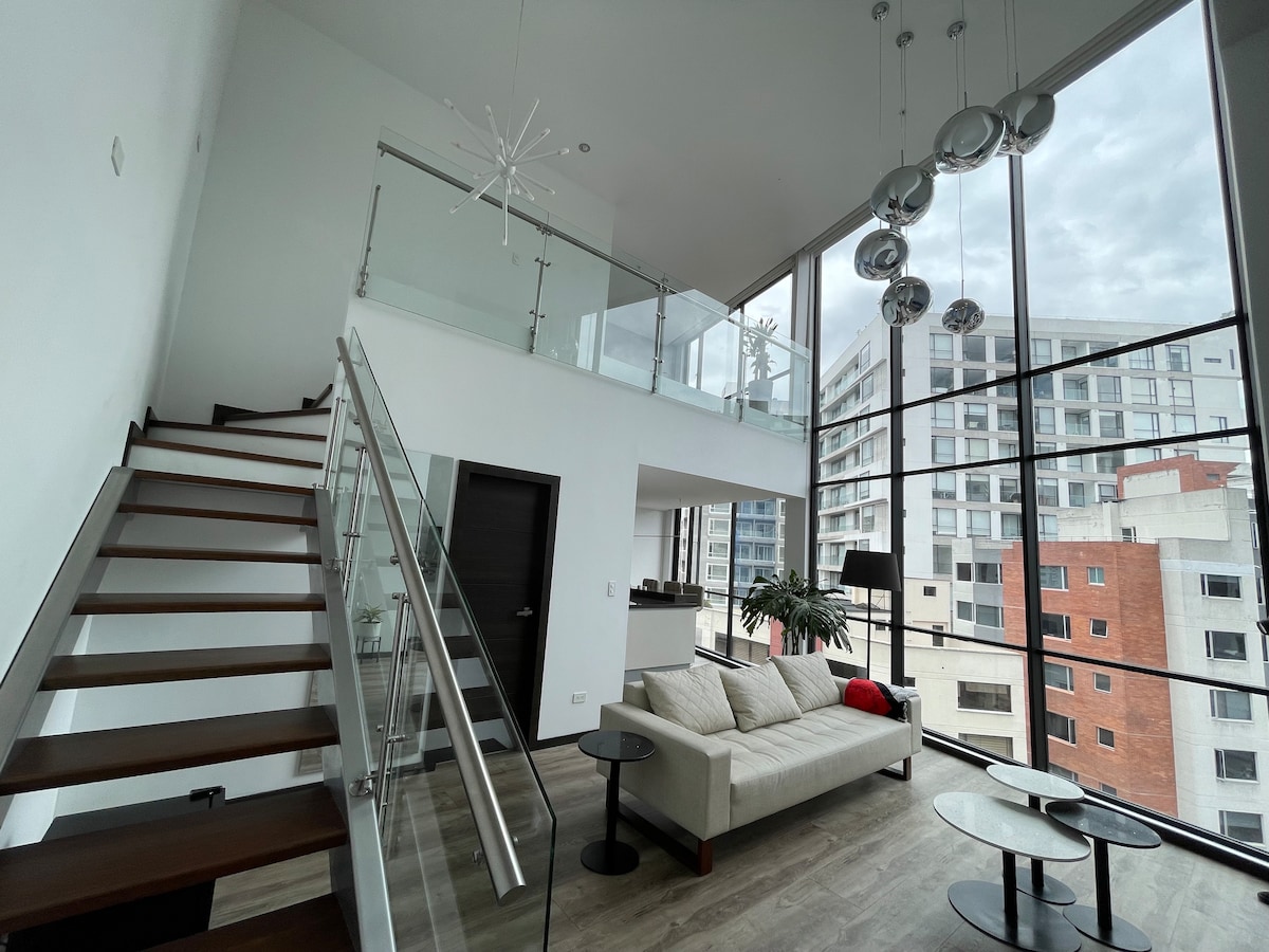 An open living area is showcased with high ceilings and large windows that provide city views. A contemporary sofa is paired with circular tables, while a staircase leads to an upper level. Modern light fixtures hang from the ceiling, adding to the elegant design.