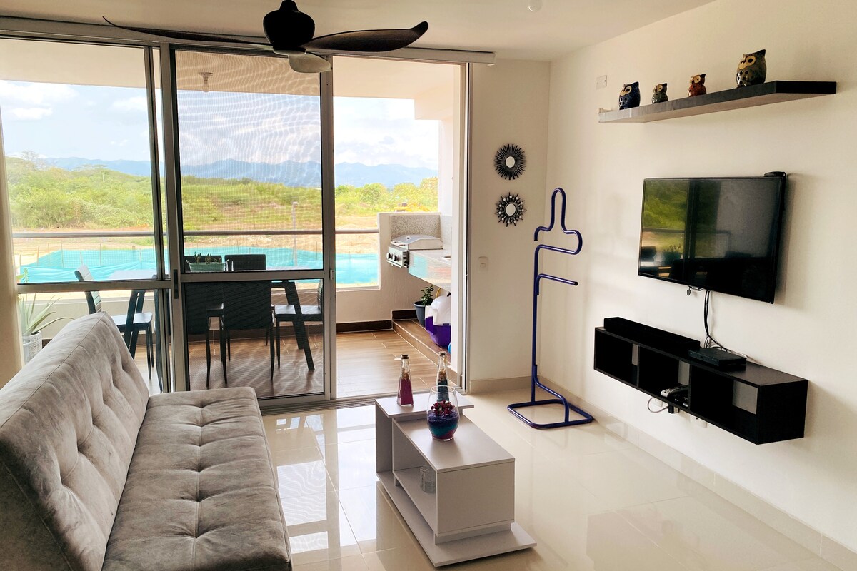A bright living area features a spacious sofa and a modern TV mounted on the wall. Large glass doors provide a view of the outdoor pool and surrounding landscape. A dining table is visible near the kitchenette, complemented by decorative elements on the shelves.