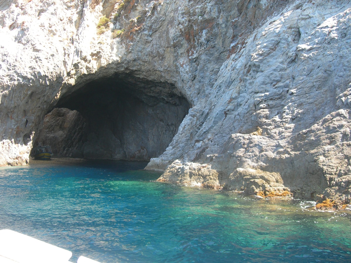 A rugged rock formation frames a coastal cave, with its dark entrance contrasting against the clear blue water. The vibrant shades of the sea blend from deep blue to turquoise, highlighting the natural beauty of the surrounding area.