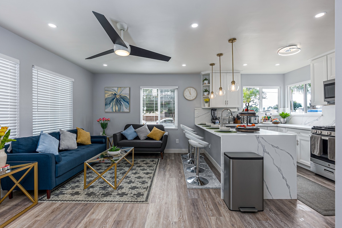 An open concept living area features a blue sofa and a dark couch, complemented by a geometric area rug. A modern kitchen showcases a marble-topped island with bar seating. Natural light fills the space through large windows adorned with white blinds.