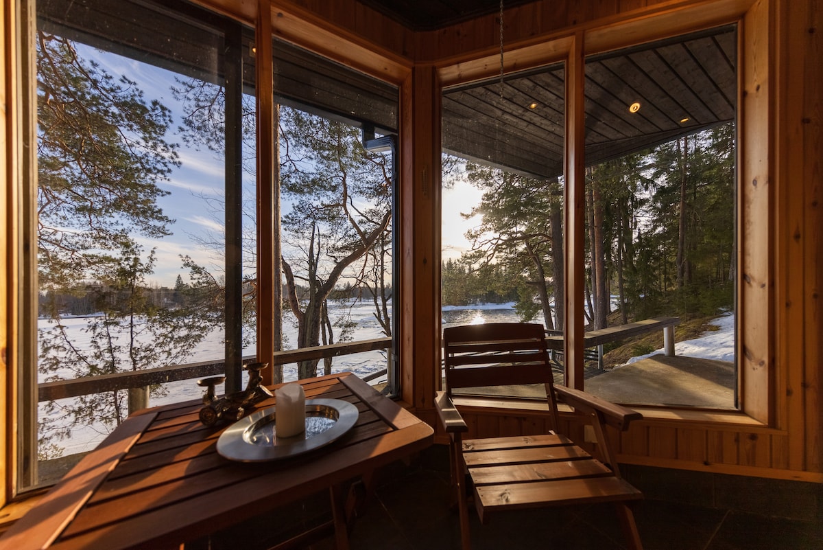 A wooden table is set near large windows, offering expansive views of the serene outdoor landscape, including trees and snow. Comfortable seating is available, and natural light fills the space, creating a peaceful retreat.