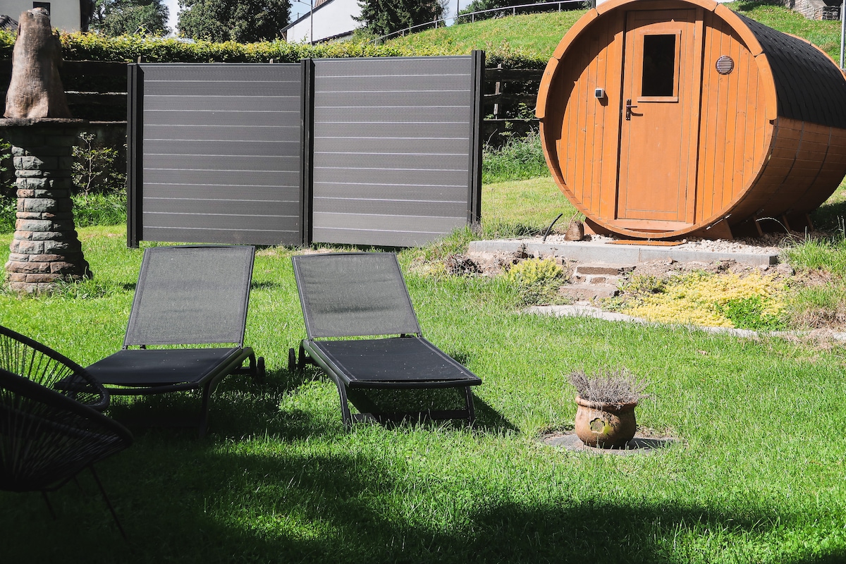 Two lounge chairs are positioned on a grassy area, offering a relaxing space in the garden. In the background, a wooden barrel sauna adds a unique element to the outdoor setting. Tall hedges provide privacy, creating a peaceful retreat.