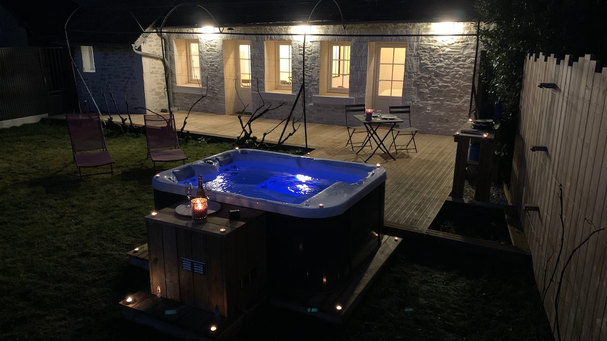 A well-lit outdoor area features a wooden terrace with a hot tub, illuminated in soft blue hues. Two lounge chairs are positioned nearby, alongside a small dining table and grill. The stone exterior of the house and large windows create a cozy ambiance, visible from the garden.