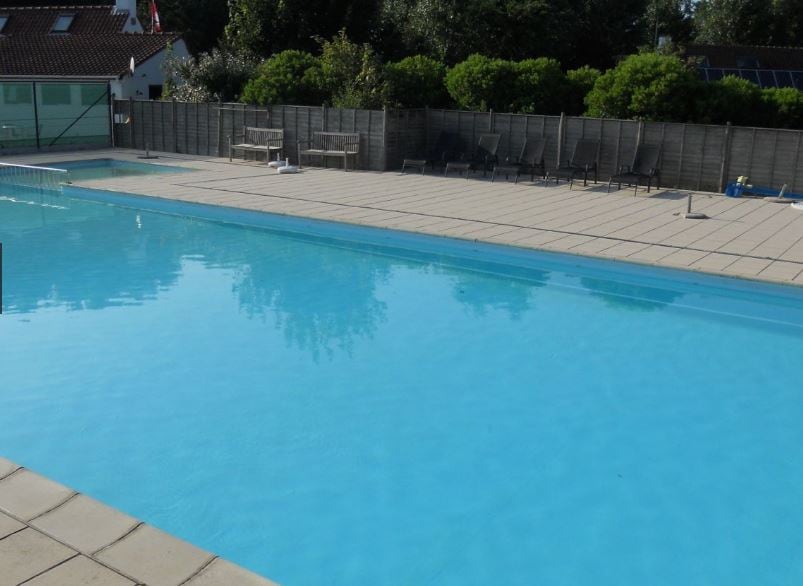 A spacious outdoor swimming pool is surrounded by tiled decking and shaded seating areas. Lush greenery provides a natural backdrop, while lounge chairs are positioned around the poolside, inviting relaxation. The clear blue water reflects the sunlight, creating a serene atmosphere.