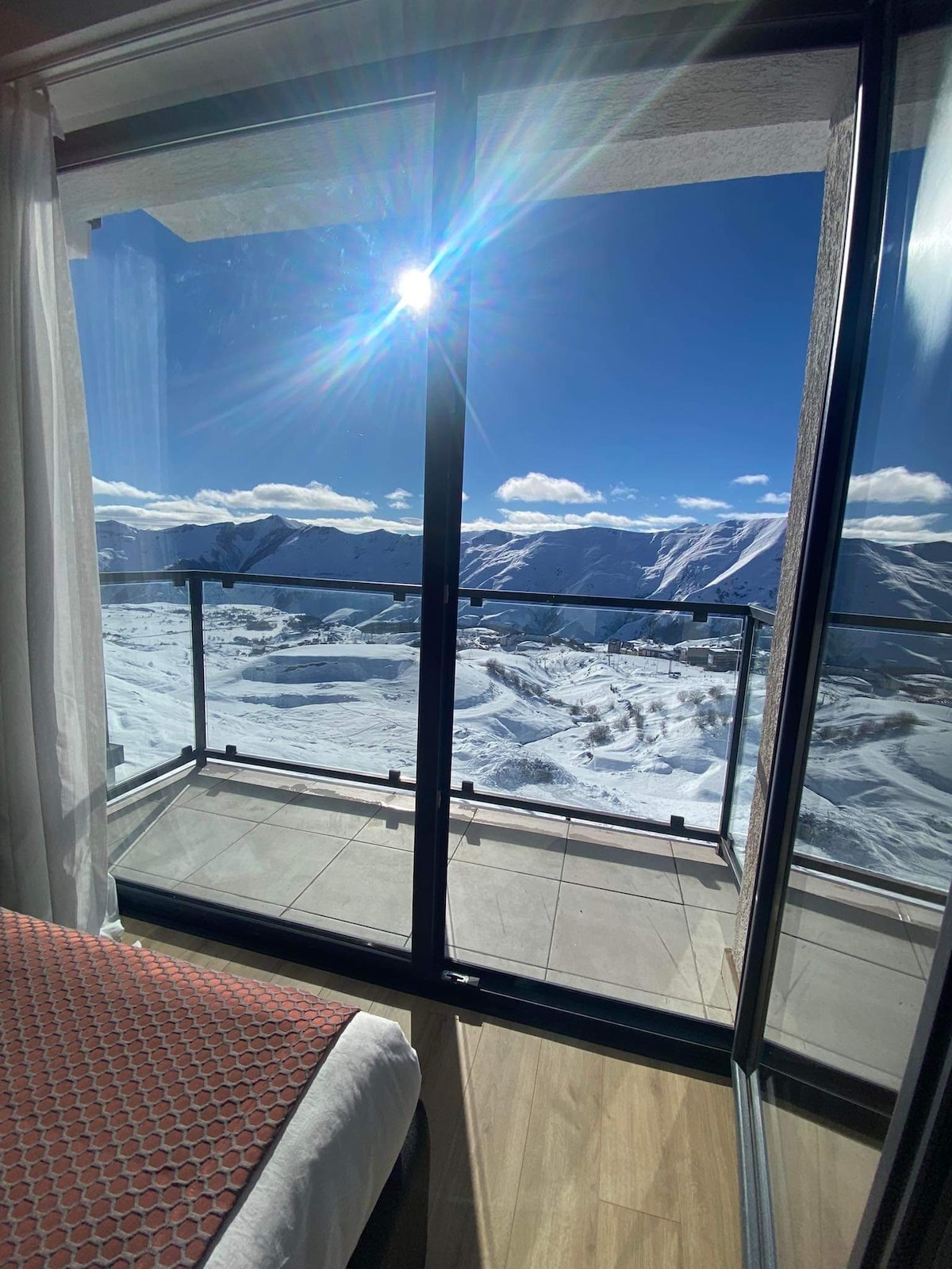 A balcony with a glass railing offers a panoramic view of snow-covered mountains under a clear blue sky. Bright sunlight reflects off the surface, illuminating the space. A portion of the spacious bedroom is visible in the foreground, featuring a patterned bedspread.