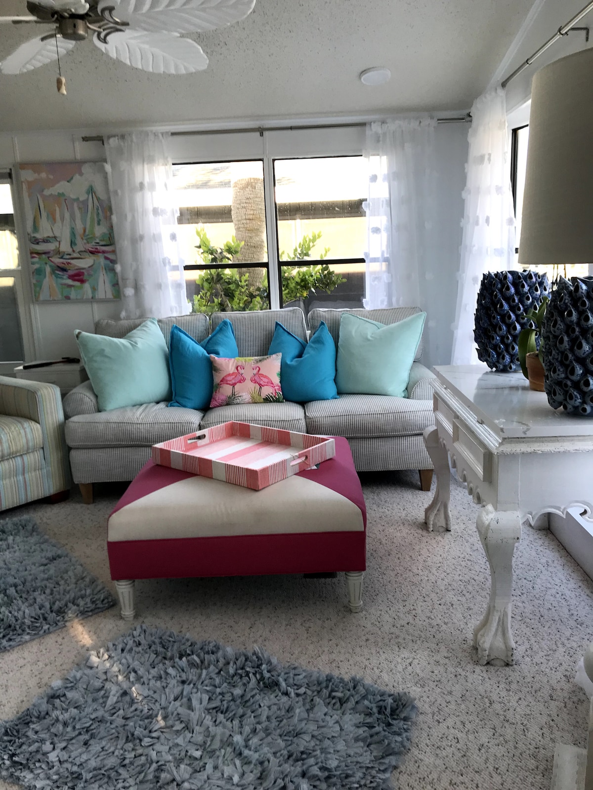 A comfortable seating area is presented with a light gray sofa adorned with vibrant blue and pink throw pillows. A pink ottoman and a decorative tray sit in front, while large windows allow natural light to fill the space, complemented by sheer white curtains.