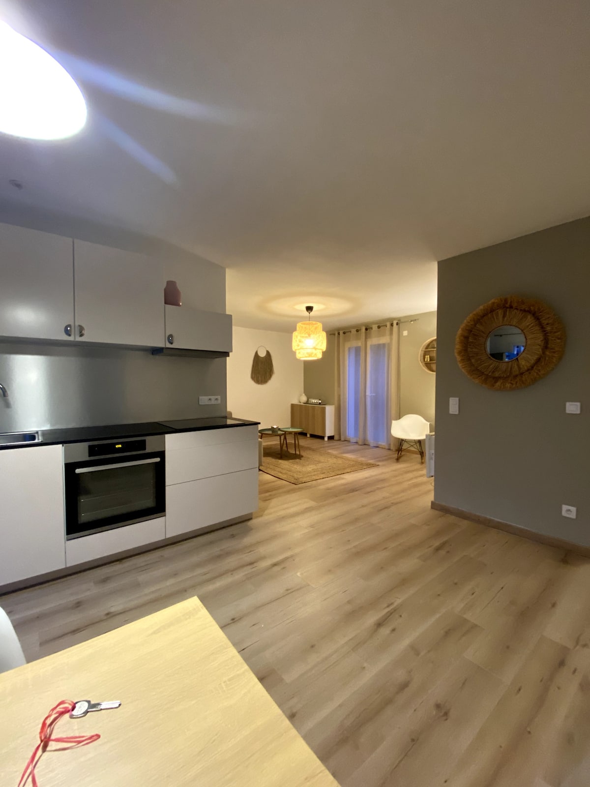 A modern kitchen area is visible, featuring sleek white cabinetry and a built-in oven. A spacious layout connects to a living area, highlighted by soft lighting and decorative elements. Wood flooring lends warmth to the overall ambience, while a circular mirror accentuates the wall.