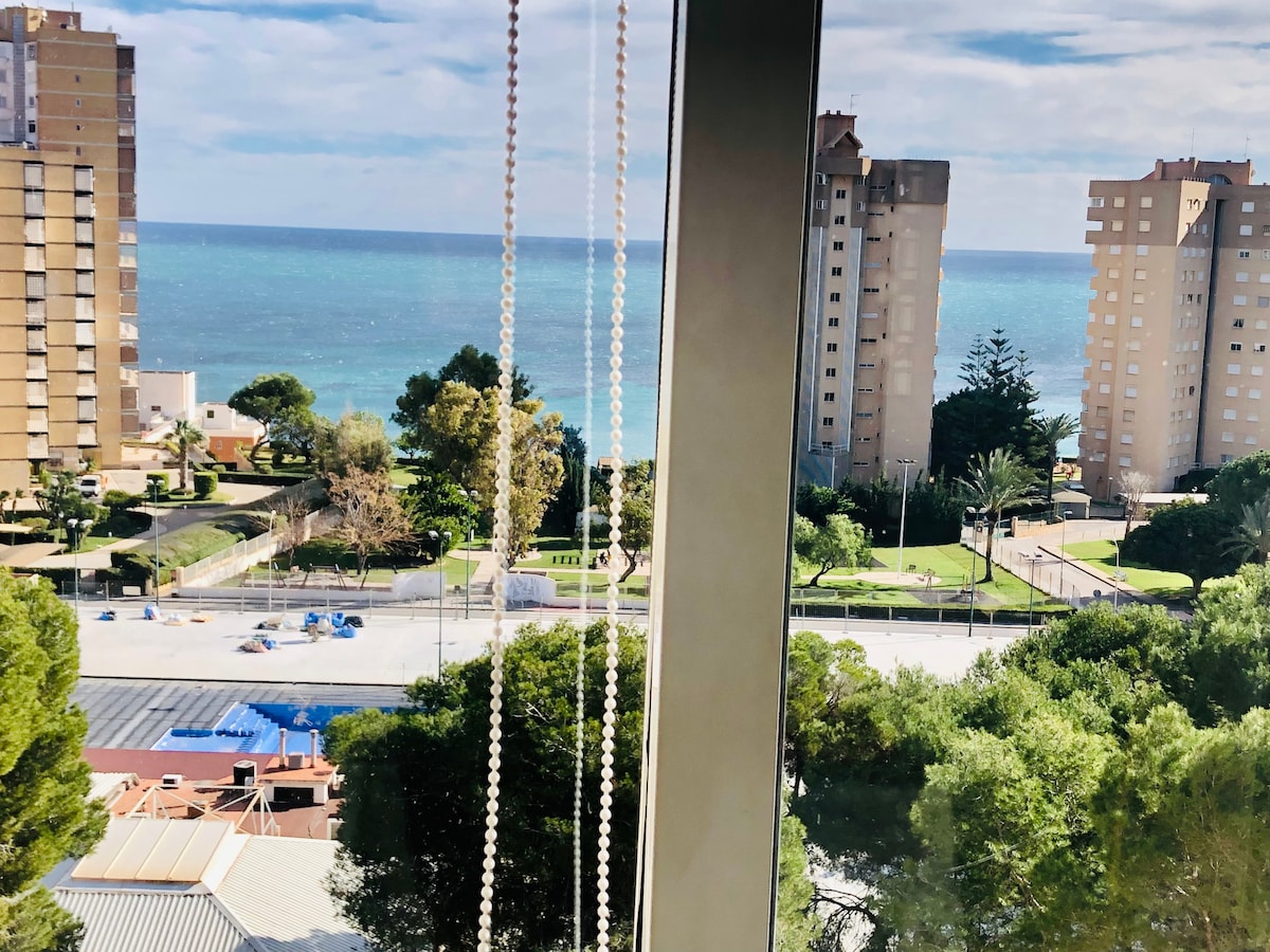 A panoramic view of the coastline is framed by large windows. The vibrant blue sea stretches into the distance, bordered by sandy shores and greenery. Nearby, modern buildings are visible alongside well-maintained gardens and pathways, enhancing the serene coastal atmosphere.