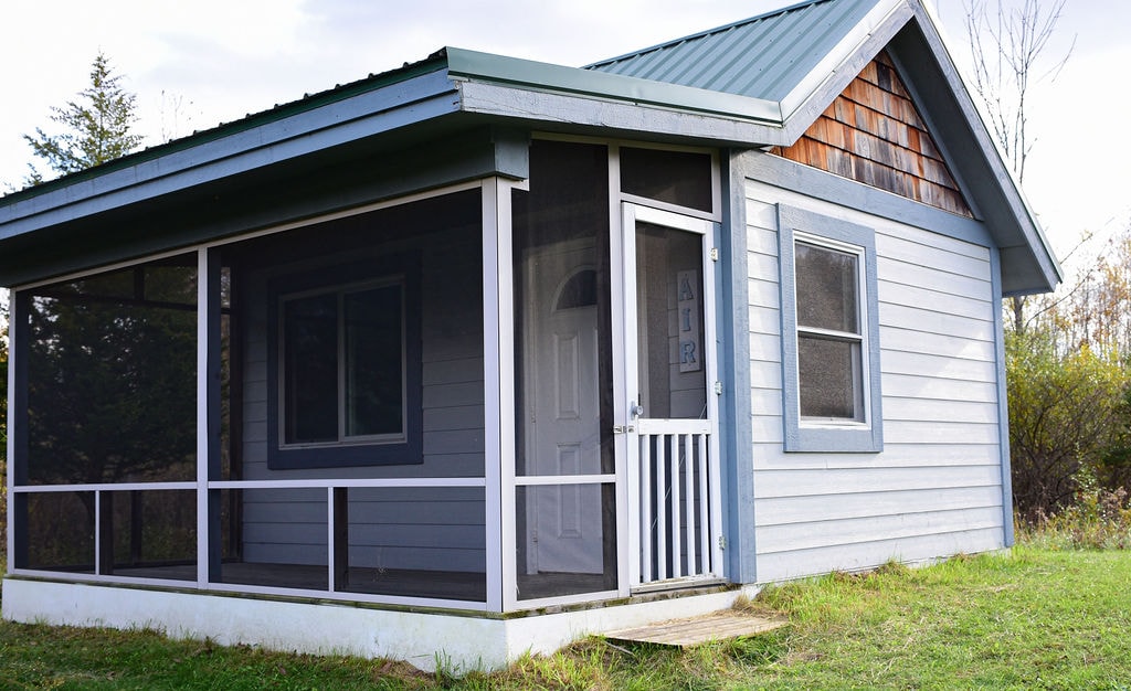 The exterior of the eco-cabin is shown, featuring a sloping green roof and a mix of blue and wood paneling. A screened porch invites guests to enjoy the surroundings, with a white door framed by a subtle front step. Grass surrounds the cabin, enhancing its natural setting.