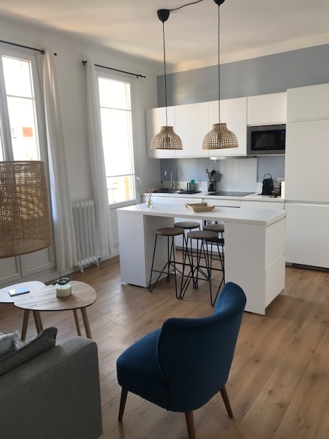A modern kitchen area is visible, featuring white cabinetry and a central island with three stools. Natural light floods the space through large windows, and two hanging pendant lights add a touch of warmth. A cozy seating area with a blue chair complements the design.